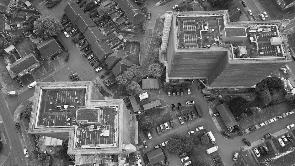 An aerial black-and-white photograph capturing a residential area during a home relocation process, featuring two multi-storey buildings with flat roofs, one on the left and one on the right. The buil