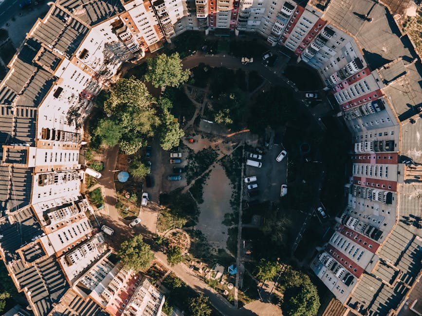 An aerial black-and-white photograph capturing a residential area during a home relocation process, featuring two multi-storey buildings with flat roofs, one on the left and one on the right. The buil