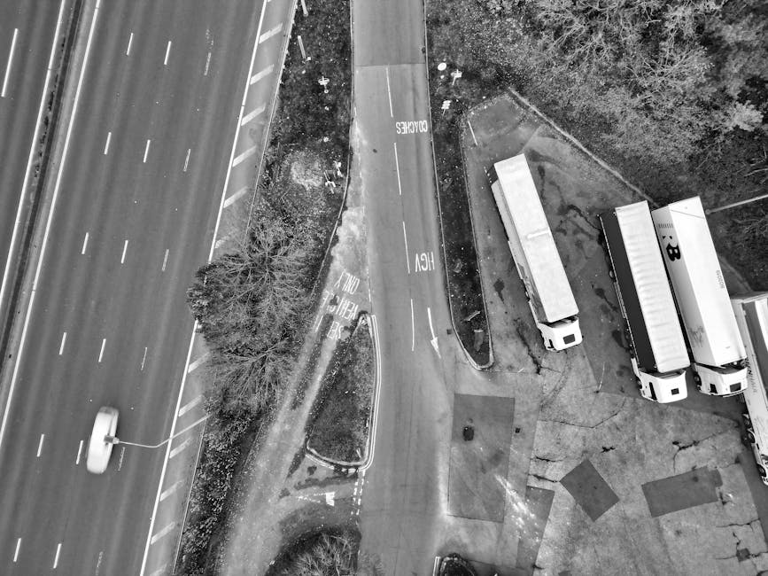 An aerial view of a multi-level highway interchange within an urban area, showing several overlapping concrete slip roads and flyovers surrounded by dense buildings, green trees, and parking lots. The